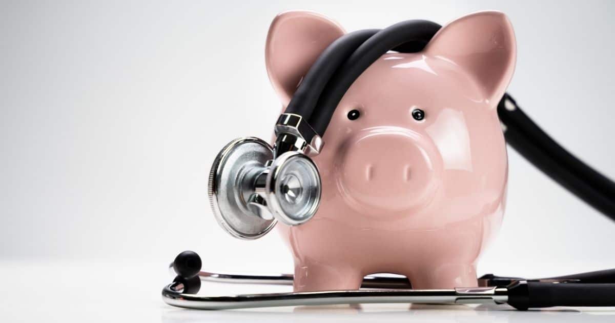 Een stethoscoop ligt gedrapeerd over een glanzend roze spaarvarken. Het varken staat op een witte tafel, tegen een witte achtergrond.