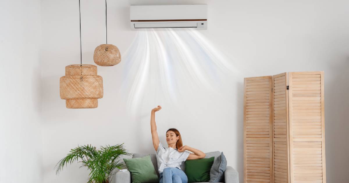 Een dame rekt zich uit onder de binnenunit van haar split airco systeem. Ze zit op een grijze bank en naast haar staat een plant.