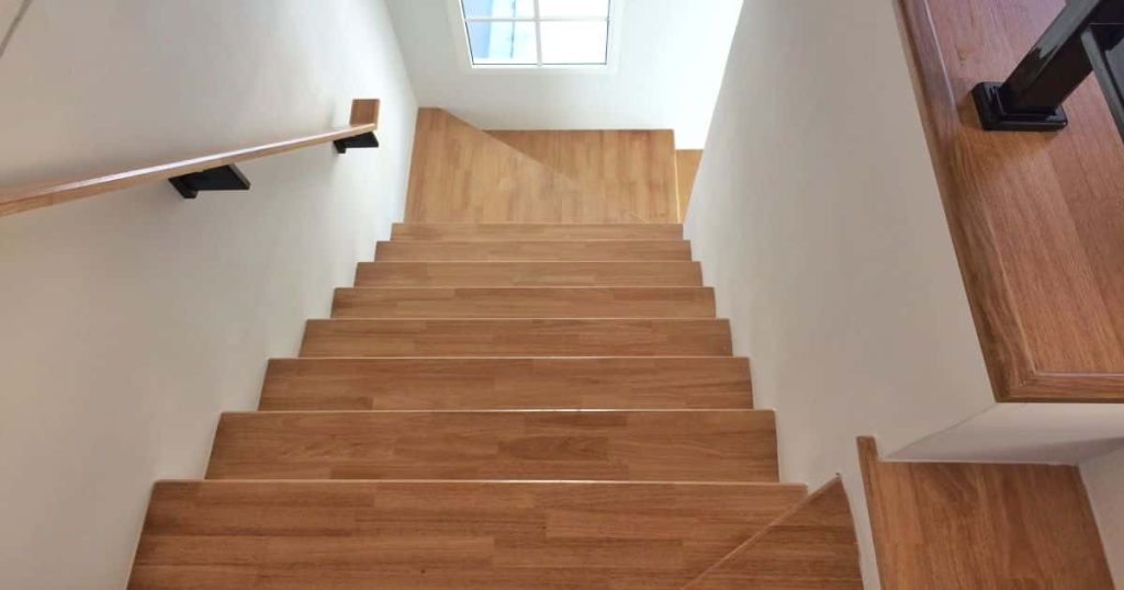 Trapgat met houten trap: de trapleuning combineert metaal/staal met hout en de muren langs de trap zijn wit geschilderd. Het raam aan het bordes beneden heeft witte kozijnen.
