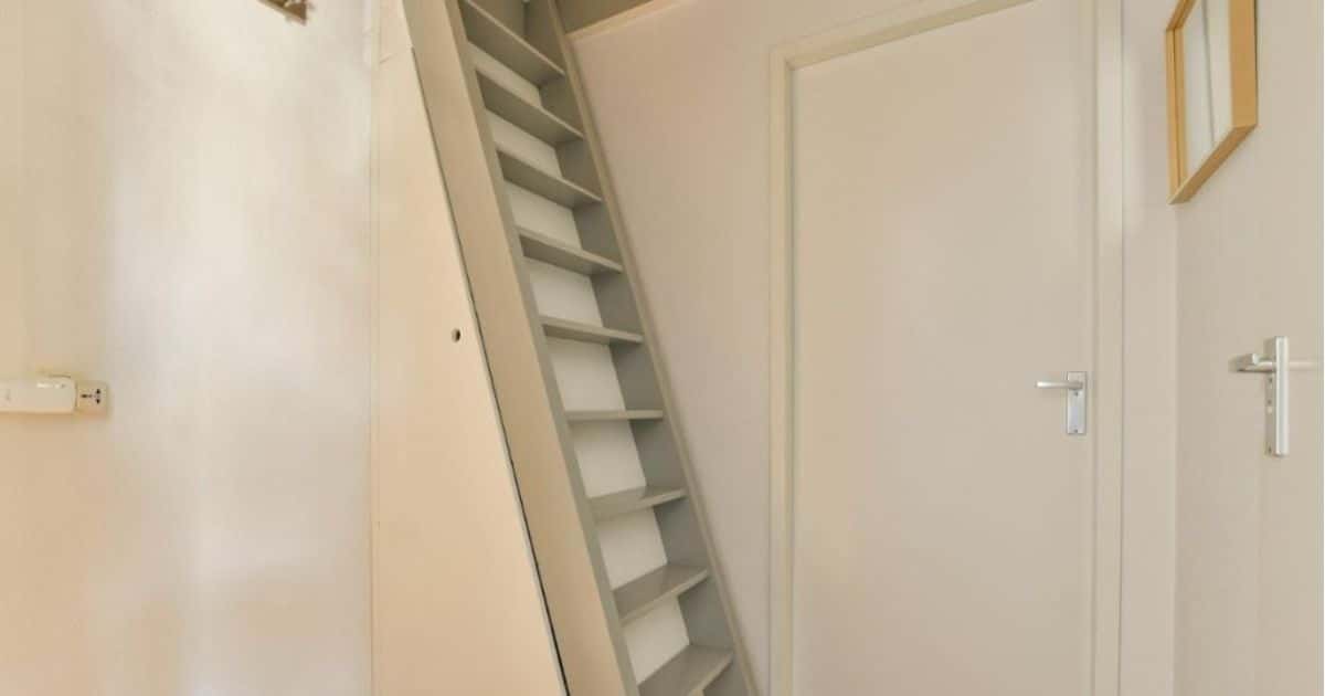 Een grijze, steile vlizotrap (trap naar de zolder) in een witte ruimte waar 2 witte deuren, gesloten, zichtbaar zijn.