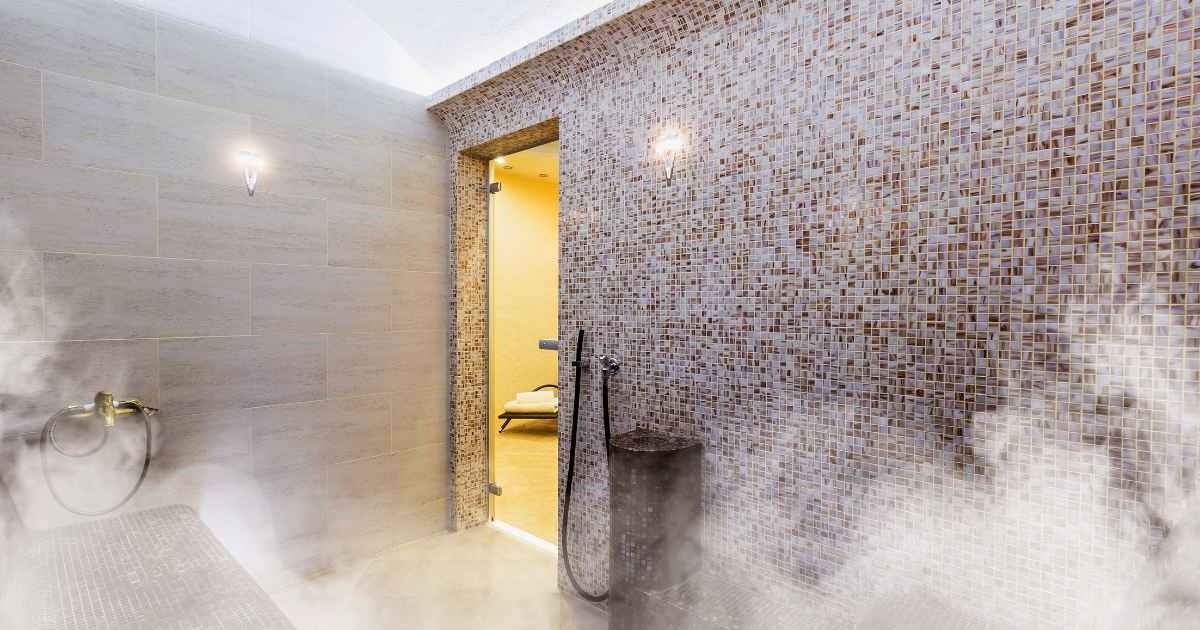 Een ruime hammam, stoombad of stoomcabine. De achterste muur is bedekt met grote tegels, de andere wand is afgewerkt met kleine mozaïektegeltjes. Er hangt veel stoom in de ruimte. Een glazen deur achterin de ruimte geeft zicht op een geel betegelde kamer.