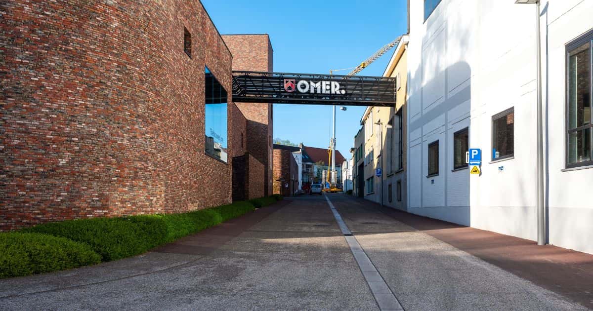 Straatzicht van de fabriek Omer in Bellegem: een brug met buizen en leidingen tussen een modern gebouw in rode bakstenen en een ouder gebouw met gele gevelsteen.