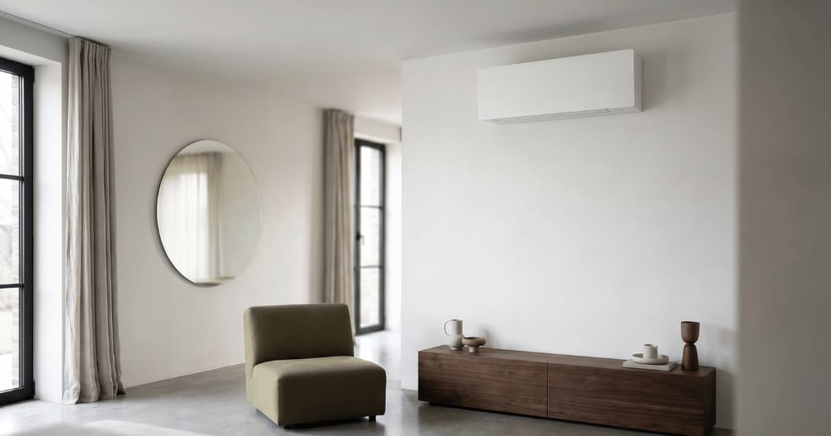 Een strakke witte airco-binnenunit hangt hoog aan de muur boven een donker notenhouten dressoir met designobjecten in een minimalistische woonkamer met gepolierde betonvloer, een olijfgroene fauteuil en een grote ronde spiegel naast hoge ramen met linnen gordijnen.