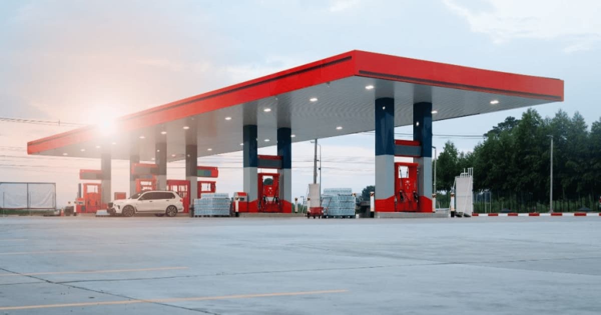 Tankstation met rood-blauwe luifel en meerdere pompen, met een geparkeerde wagen aan een van de pompen bij zonsondergang.