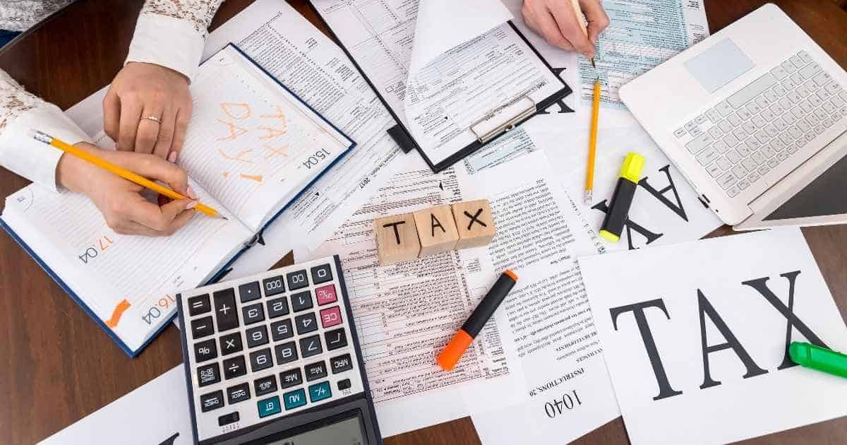 Twee mensen zitten aan een houten tafel en vullen hun belastingaangifte in. Op de tafel liggen verschillende papieren, een rekenmachine, en een groene, oranje en gele fluostift.