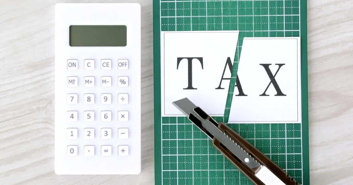 Een wit rekenmachine naast een groene snijmat met vierkantjes. Op de snijmat ligt een breekmes en een in 2 gesneden papiertje met, in hoofdletters, het woord tax (Engels voor belasting).