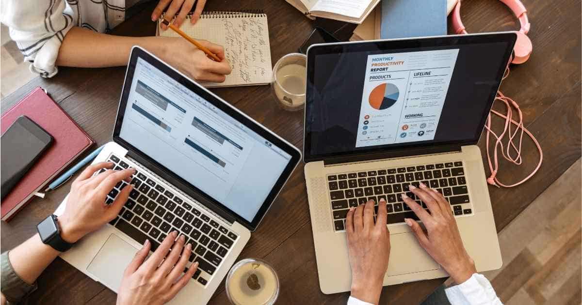 Drie mensen zitten samen aan een houten tafel. Enkel hun armen zijn zichtbaar. De persoon links vooraan typt op een laptop. De persoon rechts vooraan werkt ook aan een laptop: op het scherm is een document met cirkeldiagram geopend. Achteraan schrijft een derde persoon in een notitieboek. Op de tafel liggen boekjes, twee glazen, een roze koptelefoon en een smartphone.