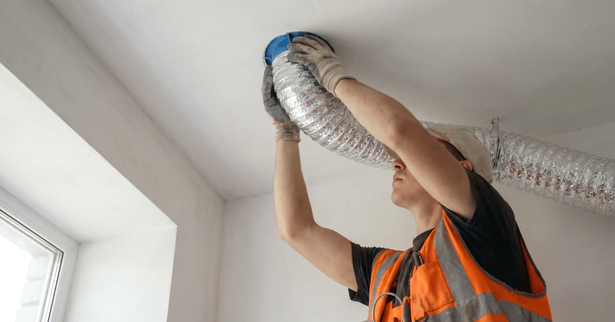 Een jonge technicus in een oranje veiligheidsvest en werkhandschoenen duwt met beide handen een aluminium flexibel kanaal op een blauw aansluitstuk tegen het witte plafond terwijl natuurlijk daglicht door het raam naar binnen valt.