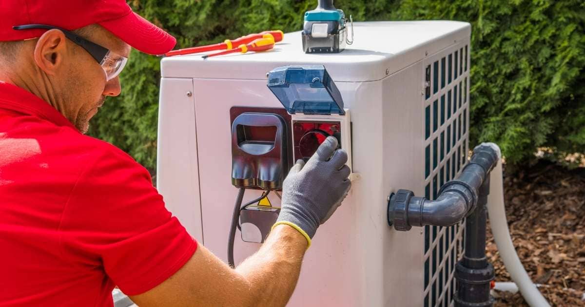 Een technicus in rood t-shirt, rode pet en zwarte veiligheidsbril werkt aan een witte warmtepomp.