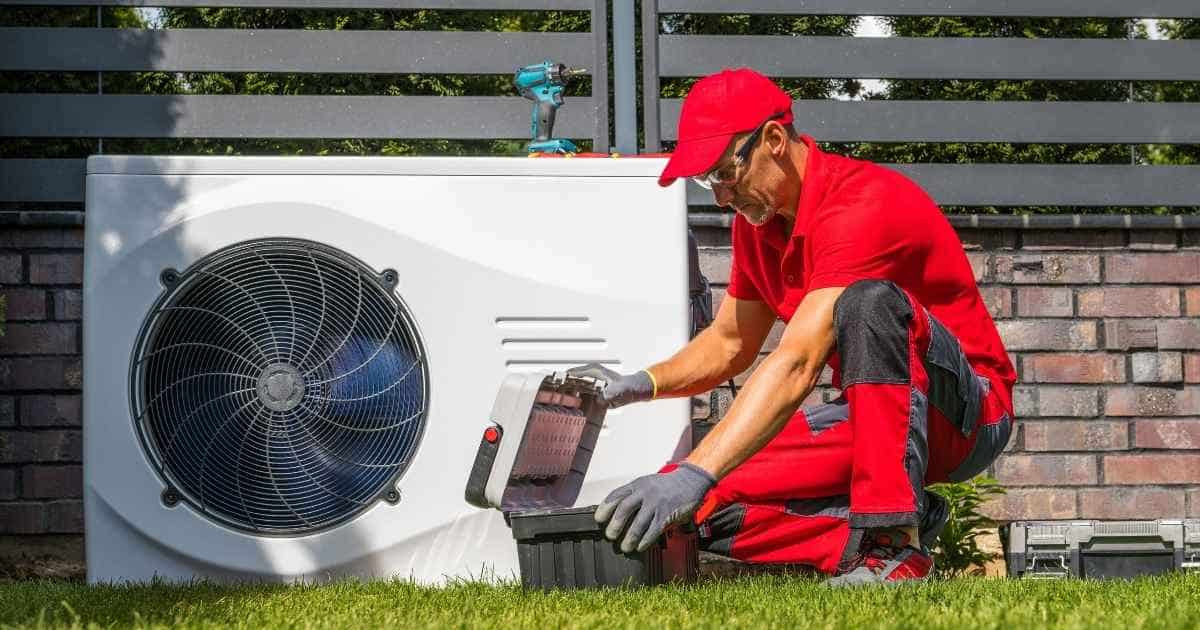 Een technicus in rode kledij werkt aan de buitenunit van een warmtepomp. Hij opent een koffer met gereedschap. Op de warmtepomp staat een blauwe boor. De technicus draagt een veiligheidsbril en werkhandschoenen.