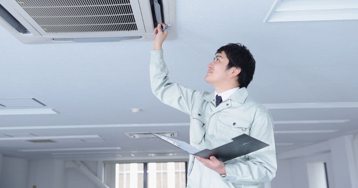 Een technicus in witte jas inspecteert een binnenunit van een airco die verwerkt werd in het plafond. Hij heeft een zwarte map vast.