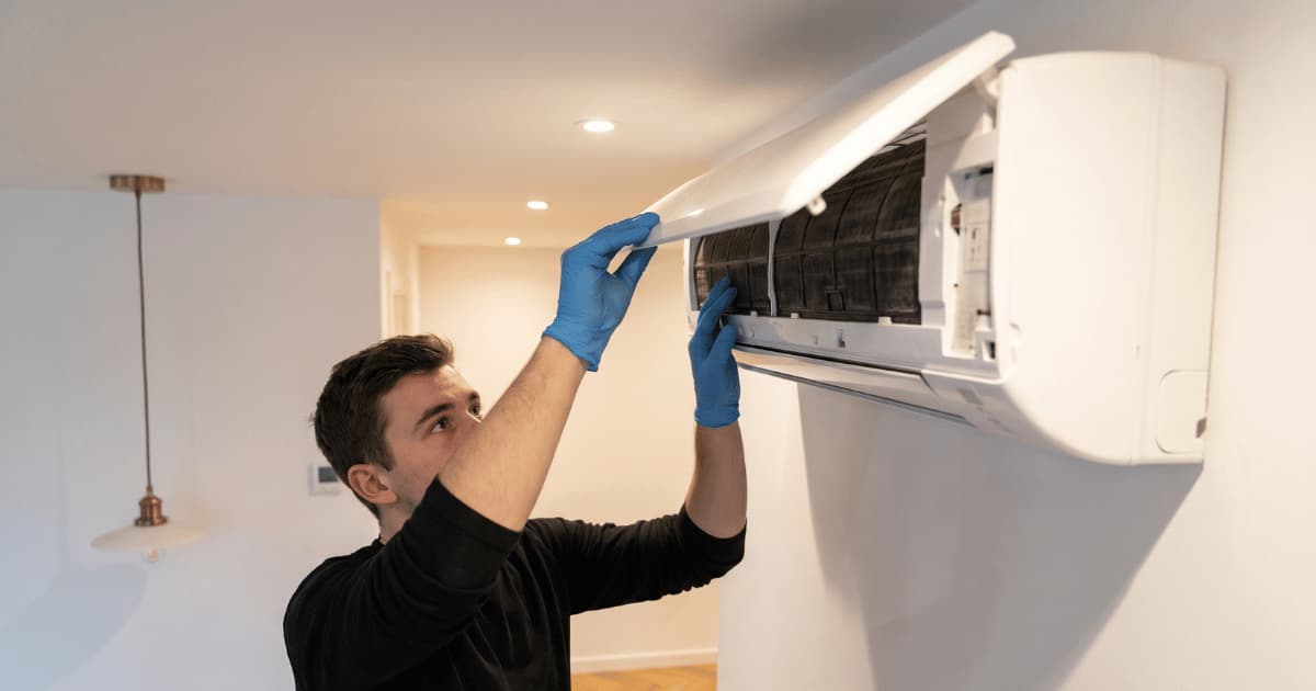 Een technicus in een zwarte trui en blauwe handschoenen opent met beide handen het frontpaneel van een witte airco binnenunit hoog aan de muur in een modern interieur met inbouwspots en koperen hanglampen.