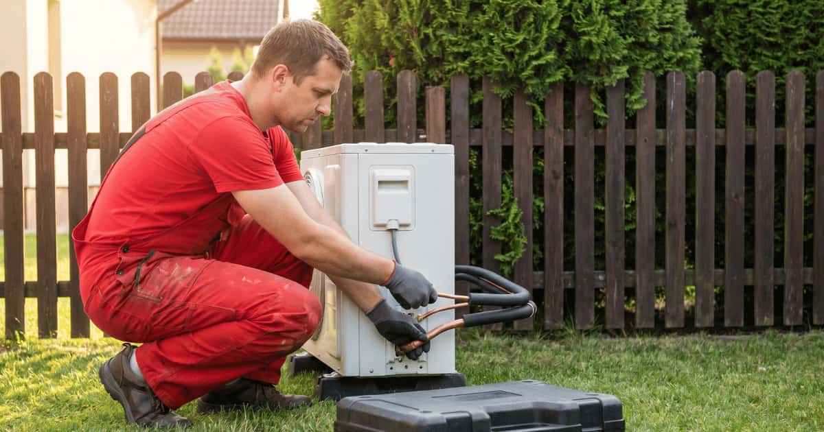 Technicus in rode werkoverall hurkt op een gazon en sluit koperen koudemiddelleidingen aan op de achterzijde van een witte warmtepomp-buitenunit naast een donkerbruin tuinhek.