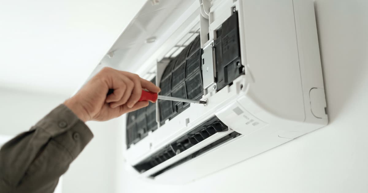 Een technicus in een kaki overhemd draait met een rode schroevendraaier aan een schroef in een geopende witte airco binnenunit met zichtbare filters en interne componenten.