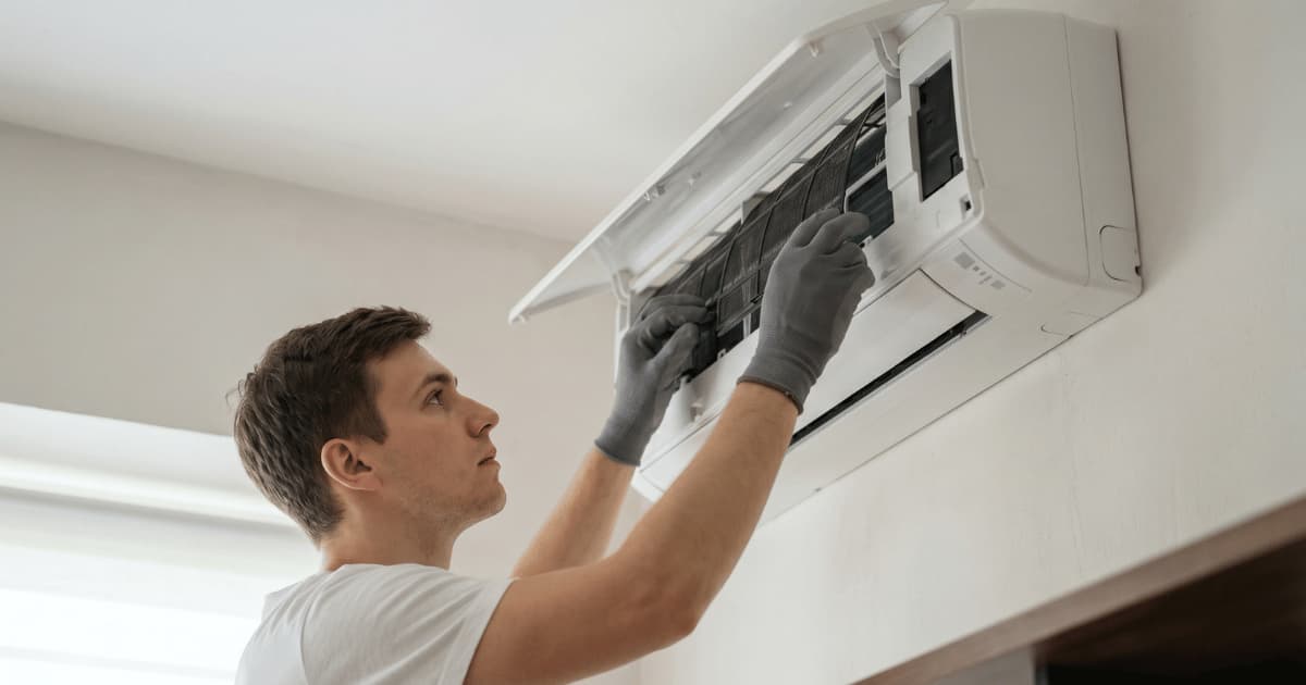 Een technicus in een wit t-shirt en grijze werkhandschoenen trekt met beide handen een filter uit een geopende witte airco-binnenunit hoog aan de muur in een lichte kamer.