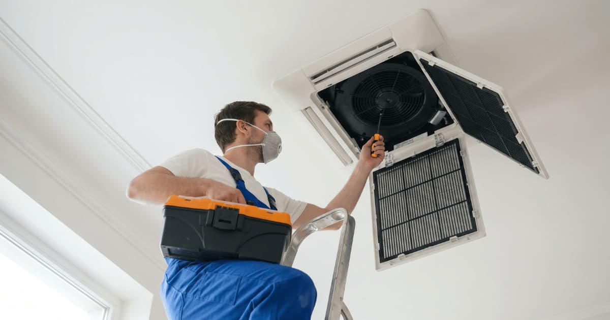 Een technieker in witte t-shirt en blauwe salopette met stofmasker staat op een aluminium trapje en gebruikt een schroevendraaier aan een geopende plafondcassette-airco waarvan de zwarte ventilator en filterroosters zichtbaar zijn.