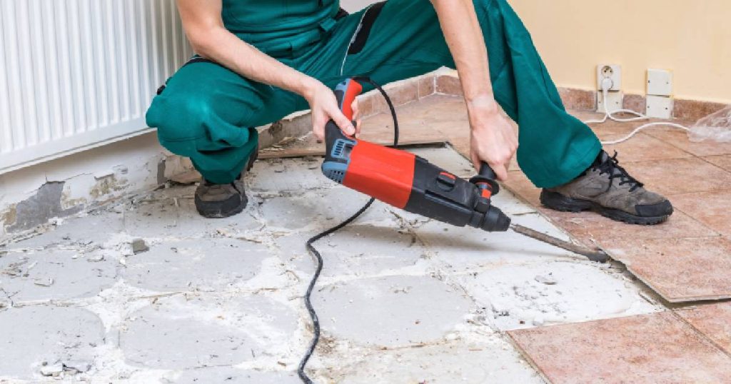 Een man gebruikt gereedschap om tegels uit te breken. Hij draagt een groene broek. Op de achtergrond links een radiator, rechts een gele muur met stopcontacten en kabels.