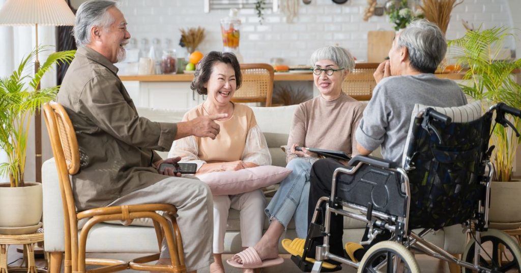 Vier ouderen zitten gezellig te praten in een woonkamer. Een van hen (een man) zit op een stoel. Een dame zit in een rolstoel, de andere twee dames op een grijs met witte zetel.