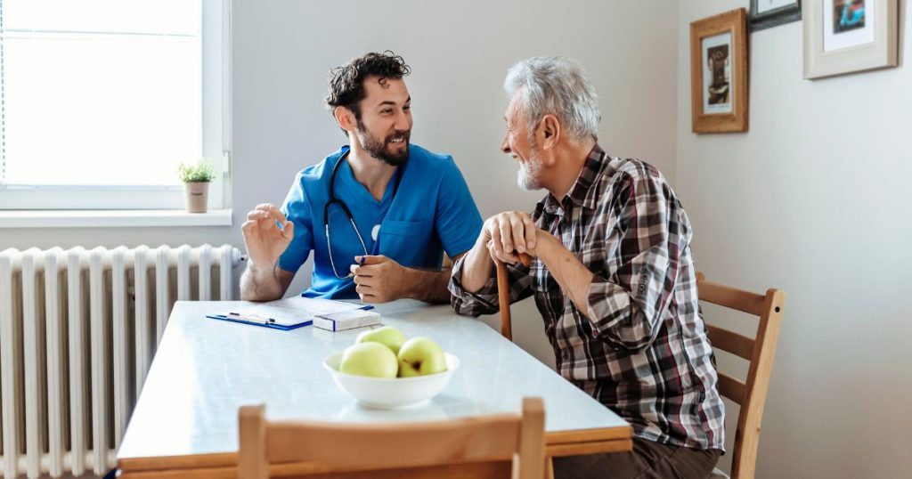 Een oudere man met wandelstok zit aan tafel met een jongere, mannelijke arts. De arts heeft een stethoscoop om de hals en draagt een blauw uniform. Op tafel liggen een klembord, schrijfgerei, en een kom met groene appels.