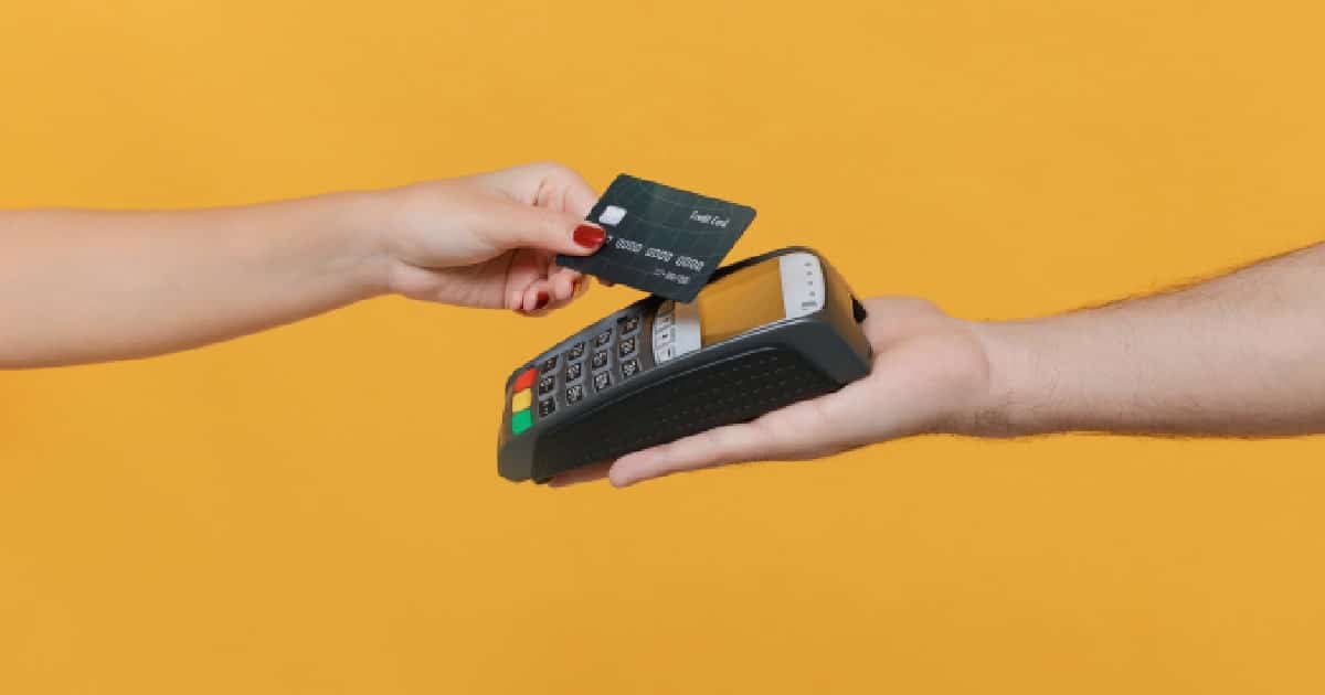 Een hand met een betaalkaart en een hand met een mobiele myPOS betaalterminal ontmoeten elkaar voor een okergele achtergrond.
