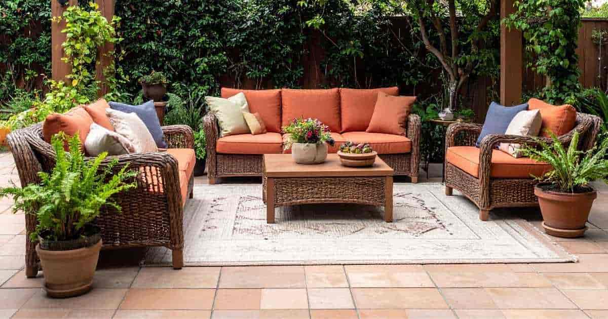 Een gezellig terras, aangelegd met oranje keramische tegels in zachte kleurschakeringen. Op het terras ligt een tapijt, met daar rond een rieten loungeset met 3 zetels en een bijzettafel. Naast de zetels staan varens in keramische potten. 