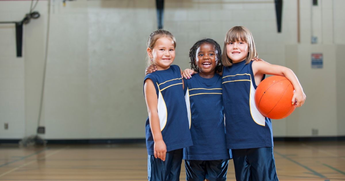 Drie meisjes in sporttenue staan op een basketbalveld, met de armen om elkaar. Het meest rechtse meisje houdt een basketbal onder haar arm.