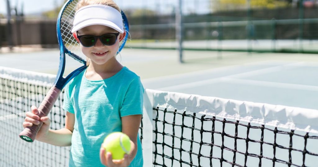 Een jong meisje houdt een tennisbal voor zich en steunt met een tennisracket tegen haar schouder. Ze staat voor een tennisnet, op een tennisveld. Ze draagt een zonnebril en tennispet.