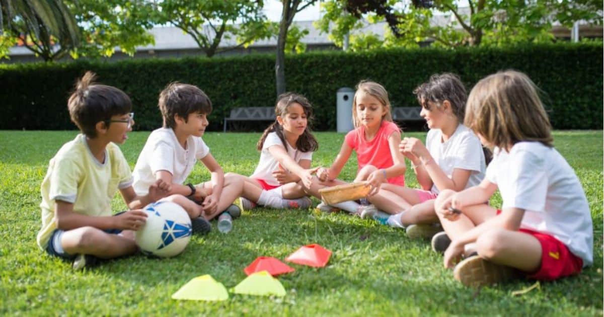 Een groep kinderen speelt een spelletje terwijl ze in een kring op een grasveld zitten.