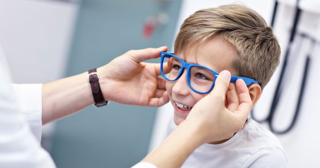 Een jongen krijgt een blauw brilmontuur aangemeten door 2 vrouwelijke handen.