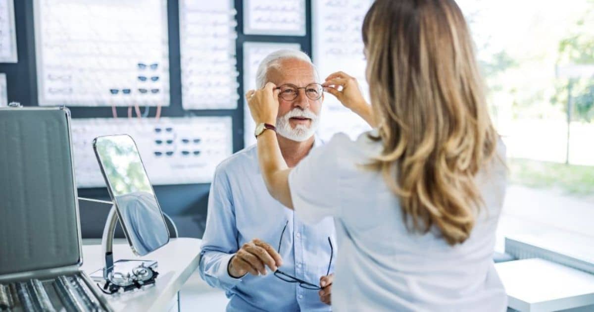Een oudere man met wit haar en een witte baard past een nieuw brilmontuur. Hij wordt geholpen door de opticien.
