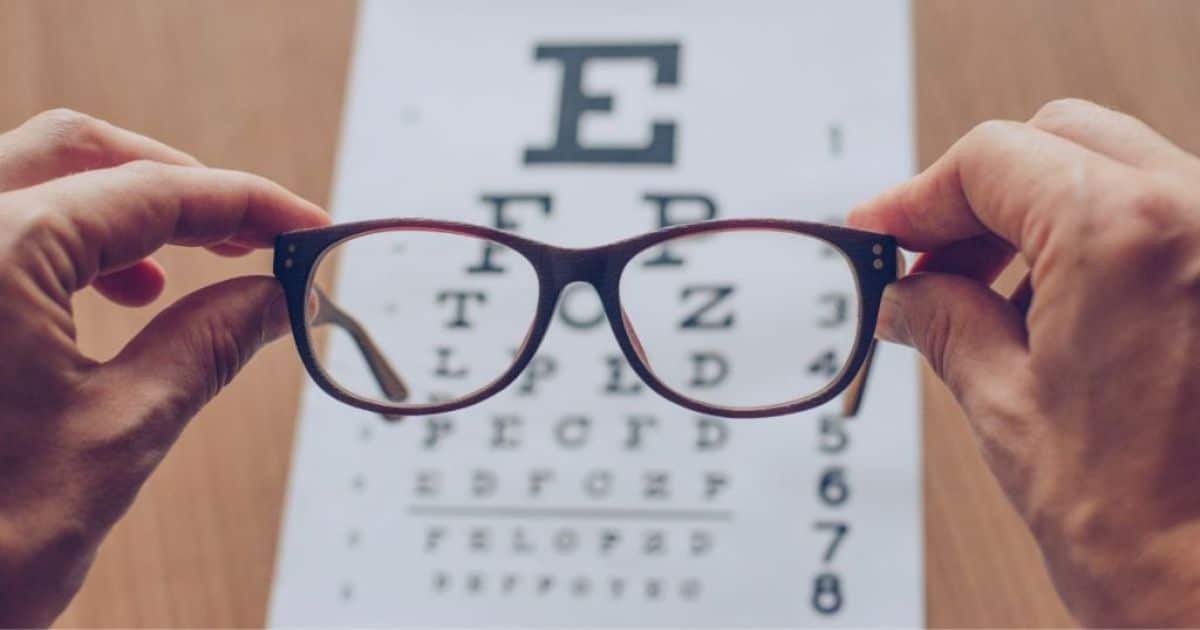 2 Handen houden een bruin brilmontuur vast. Op de achtergrond een poster met cijfers en letters van het alfabet in uiteenlopende groottes.