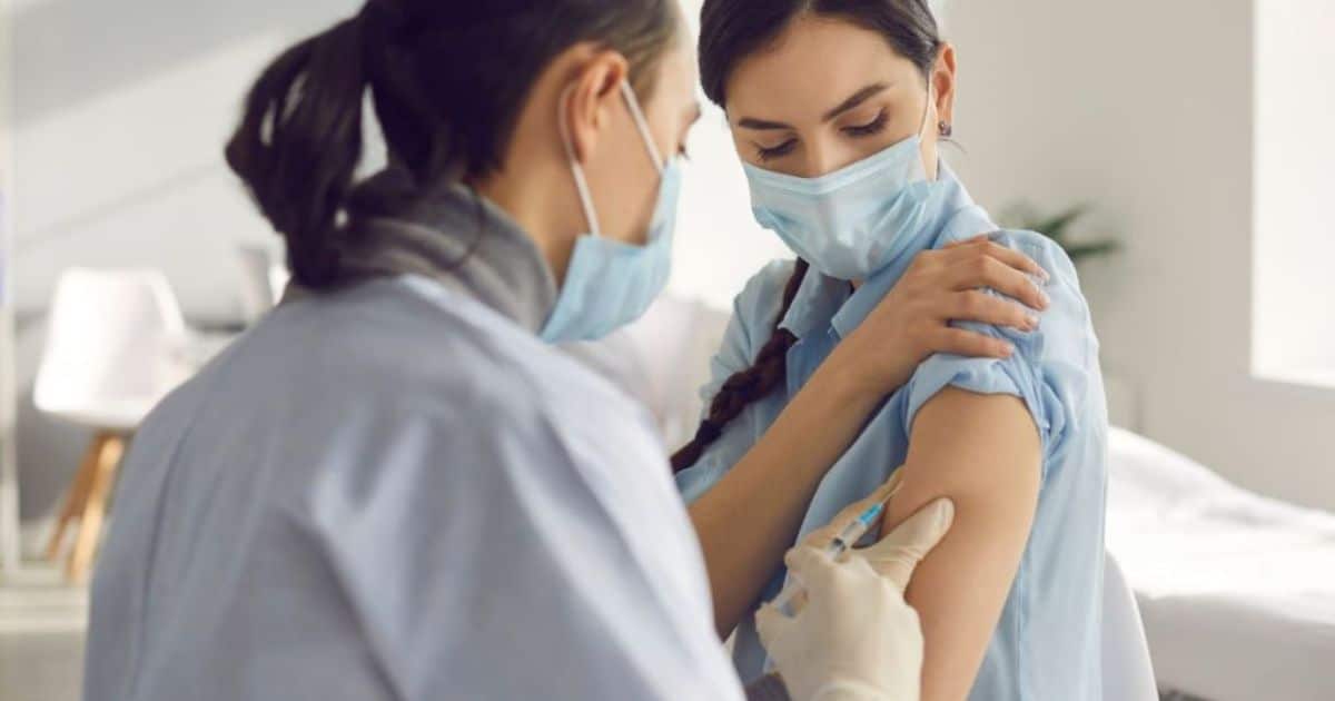 Een arts injecteert het griepvaccin in de arm van een jong persoon. Ze dragen allebei een blauw hemd en blauw mondmasker.