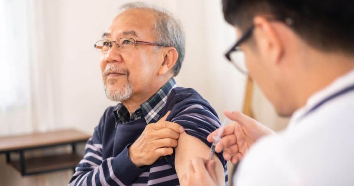 Een arts met bril geeft het griepvaccin aan een oudere man met een bril, gestreepte trui over een geruit hemd, en een baardje.