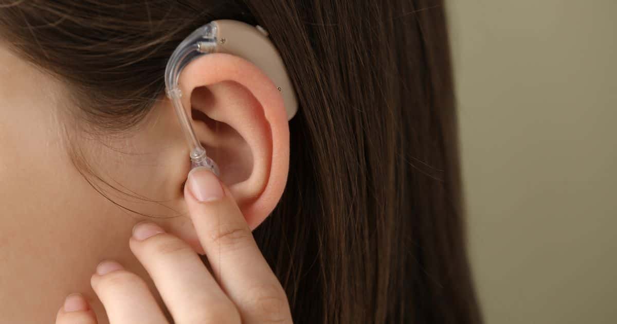Het oor van een dame met een hoorapparaat en bruin haar. Ze houdt haar wijsvinger tegen het hoorapparaat.