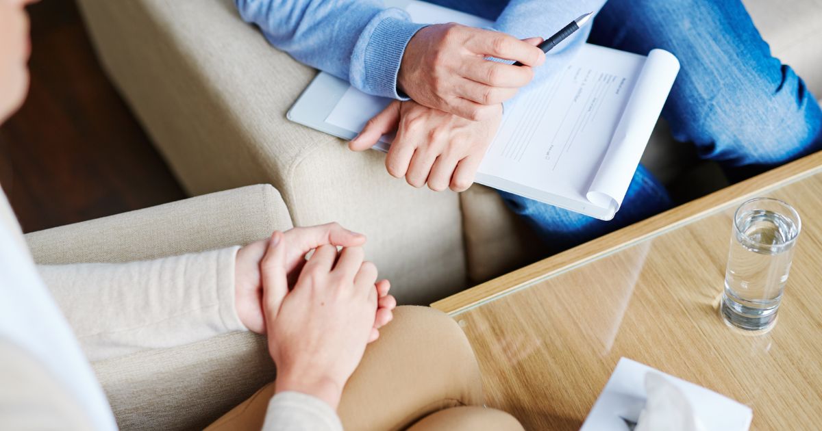Bovenaanzicht van de handen van enerzijds een patiënt en anderzijds een klinisch psycholoog of therapeut met klembord en pen. Tussen therapeut en patiënt staat een houten tafel met glazen oppervlak, waarop een glas water en doos zakdoeken staan.