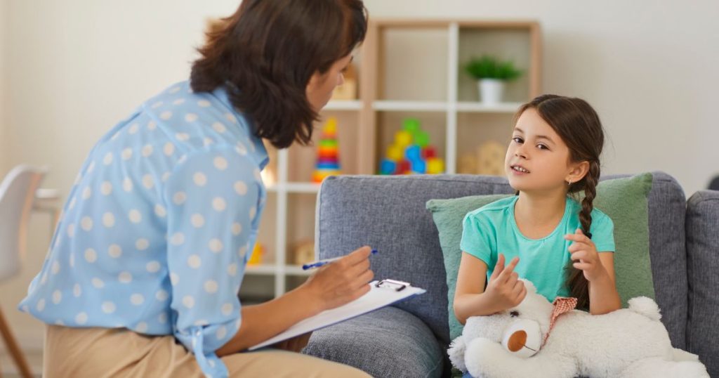 Een kinderpsychologe en een jonge patiënt. De patiënt houdt een witte beer op haar schoot. De psychologe heeft een klembord en een pen vast.