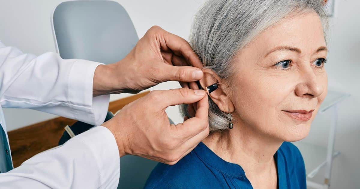 Een audicien plaatst een hoorapparaat in het oor van een dame met grijs haar en een blauw t-shirt.