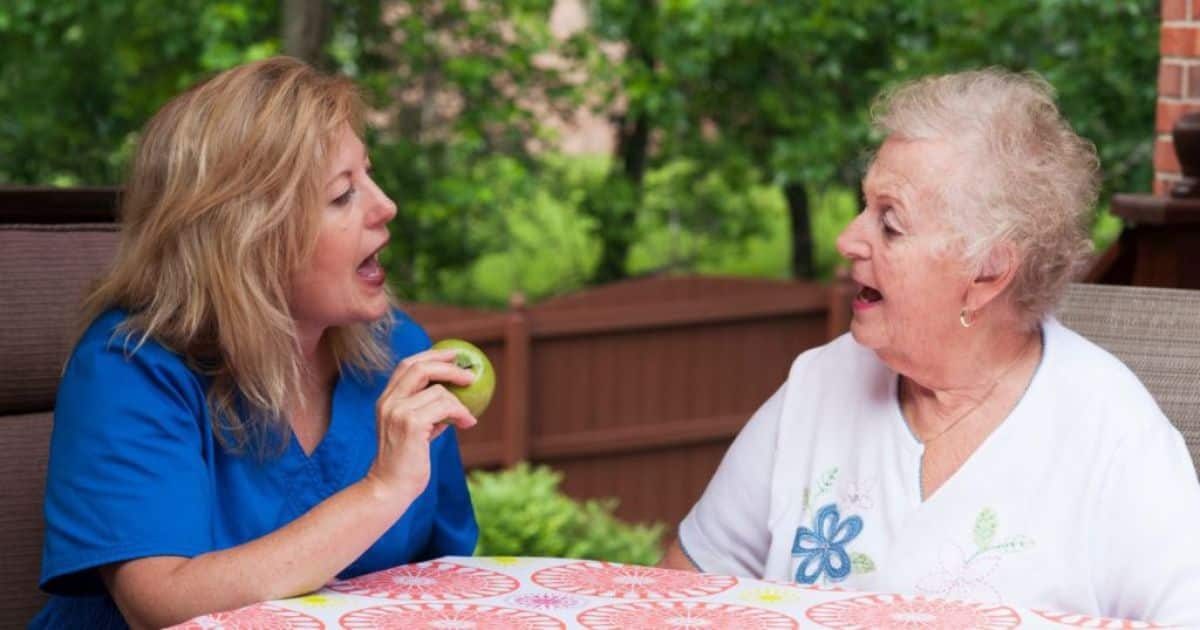 Een logopedist opent haar mond wijd terwijl ze een appel vastheeft. Een oudere dame, haar patiënt, imiteert.