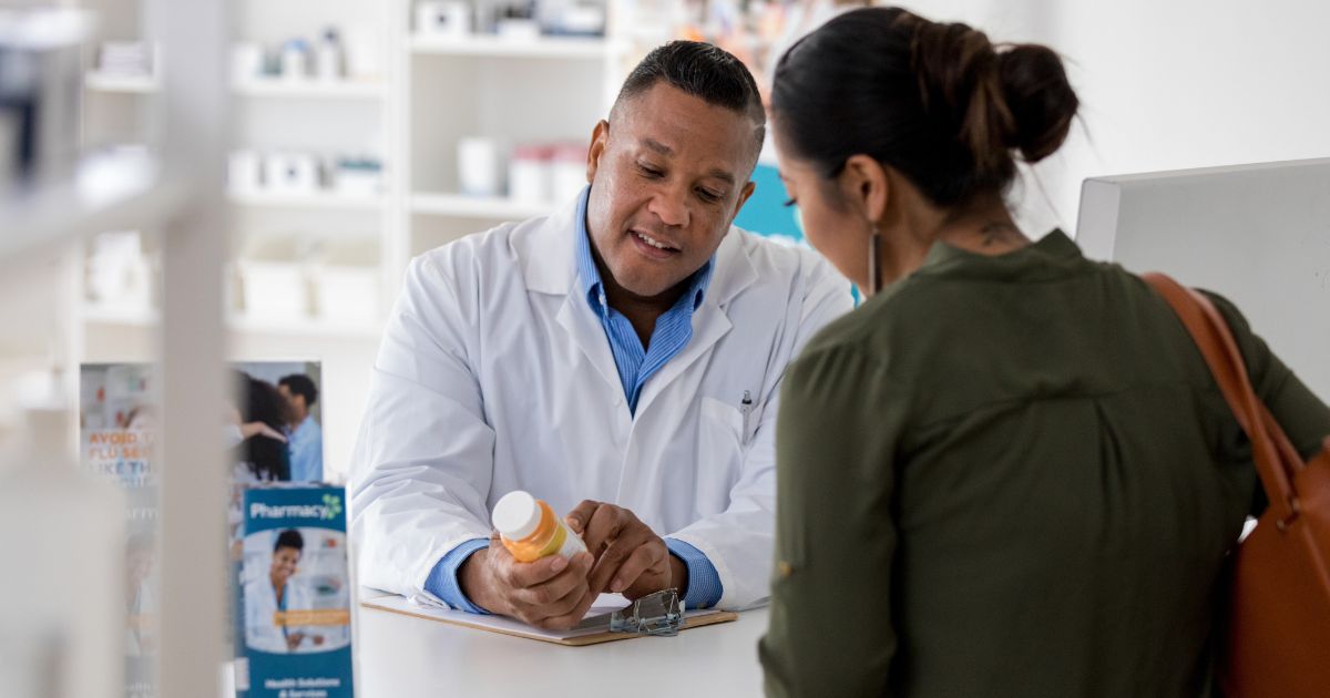 Een apotheker geeft uitleg bij een potje medicijnen aan zijn klant: een dame met bruin haar, in een groen hemd met een leren tas rond de schouder. De twee staan aan de toonbank van de apotheek, waarop een klembord ligt en enkele brochures staan.
