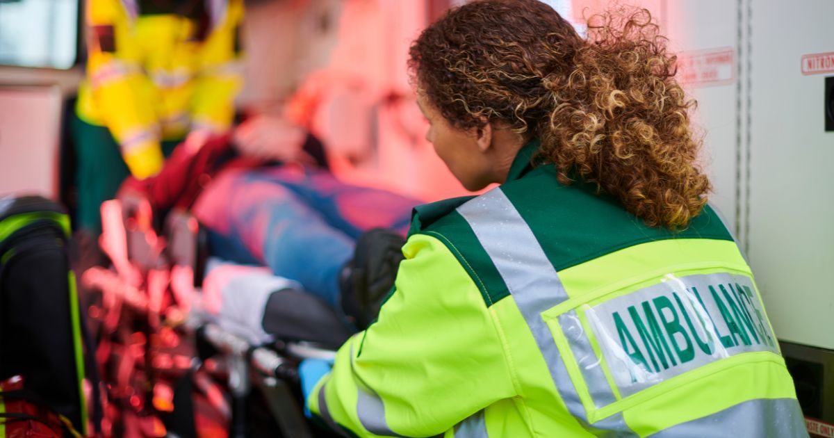 Een ambulancier met fluohesje helpt een patiënt op een brancard de ambulance in te dragen.