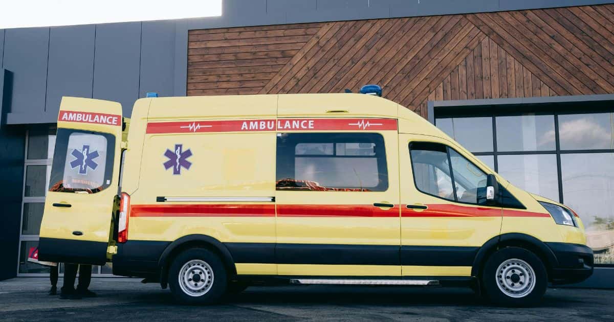 Een gele ziekenwagen met een rode streep in het midden en bovenaan, en met het opschrift 'ambulance' staat voor een publiek gebouw. De achterdeuren van de ziekenwagen staan open.