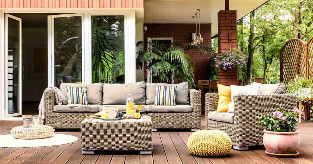 Een gezellig terras met houten plankenvloer tegen een bakstenen gevel met grote ramen en een glazen deur. Op het terras staat een rieten loungeset met 2 zetels en een lage tafel. 