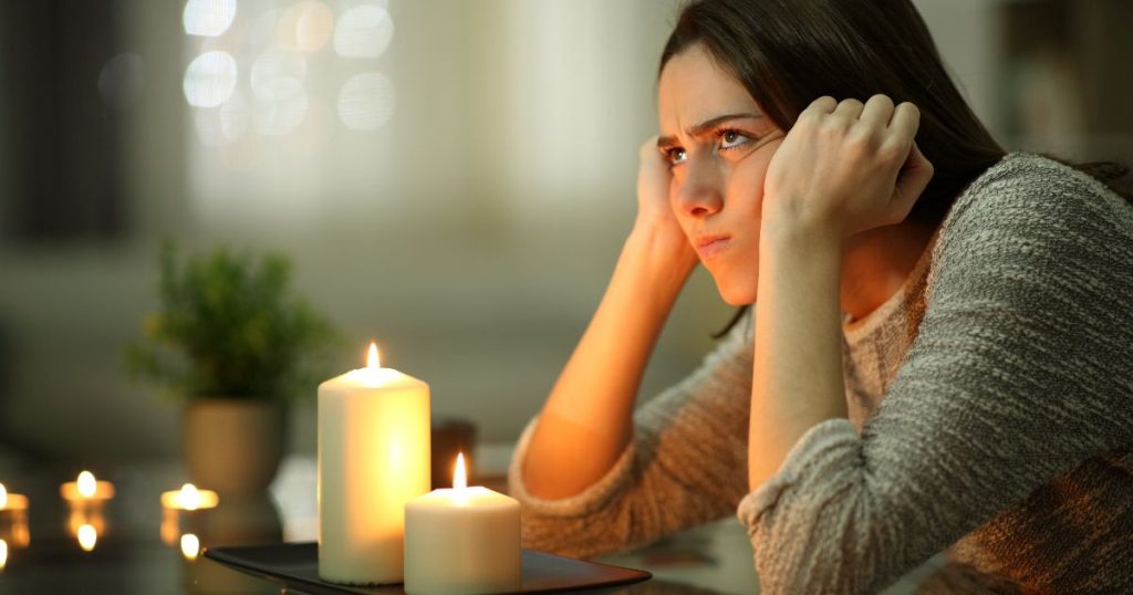 Een jonge vrouw met lang bruin haar en een grijze trui zit boos aan een tafel met kaarsen tijdens een stroomuitval. Ze houdt haar handen tegen haar gezicht en fronst.