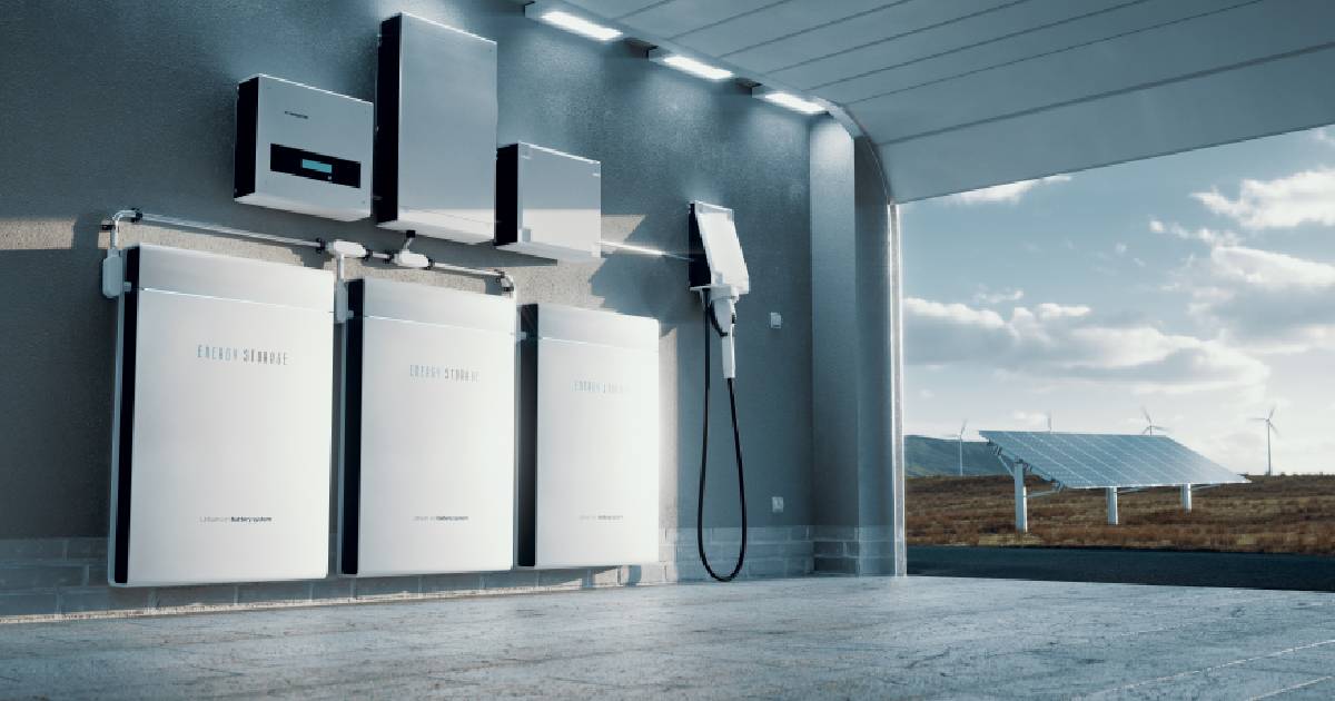 Een thuisbatterij installatie in een garagebox, rechts ervan hangt ook een laadpaal aan de muur. Op de achtergrond zie je ook zonnepanelen en windmolens.