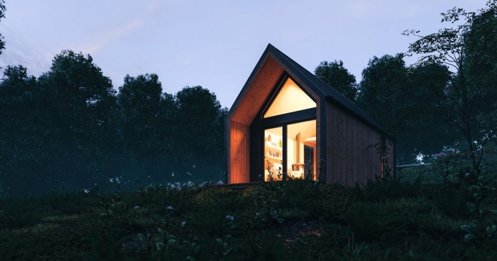 Een kleine houten woning op een heuvel- of bergflank in een bosrijke omgeving. Op het hellend dak liggen zonnepanelen. Het schemert, daarom brandt de verlichting. Het resultaat van een thuisaccu die de opgeslagen zonne-energie nu omzet in elektriciteit?