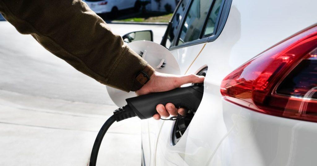 Een hand houdt een laadkabel tegen de laadpoort van een witte elektrische auto.