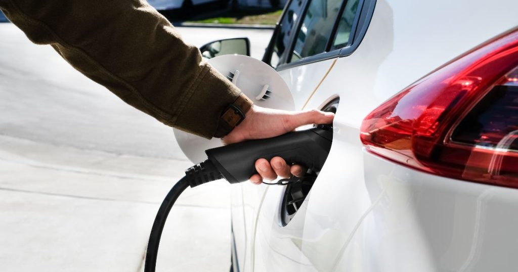 Een hand in bruine mouw steekt een oplader in een witte elektrische wagen.