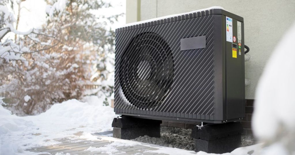 Een warmtepomp geïnstalleerd tegen een lichtgekleurde gevel, in een besneeuwde tuin.
