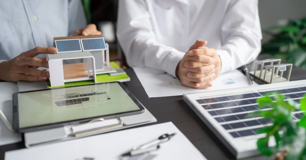 Twee mensen aan tafel: de ene heeft de handen in elkaar bovenop een blad, de ander houdt beide handen op een kleine maquette van een energiezuinige woning met zonnepanelen. Voor de maquette liggen een tablet (bovenop een klembord) en een notitieboek.