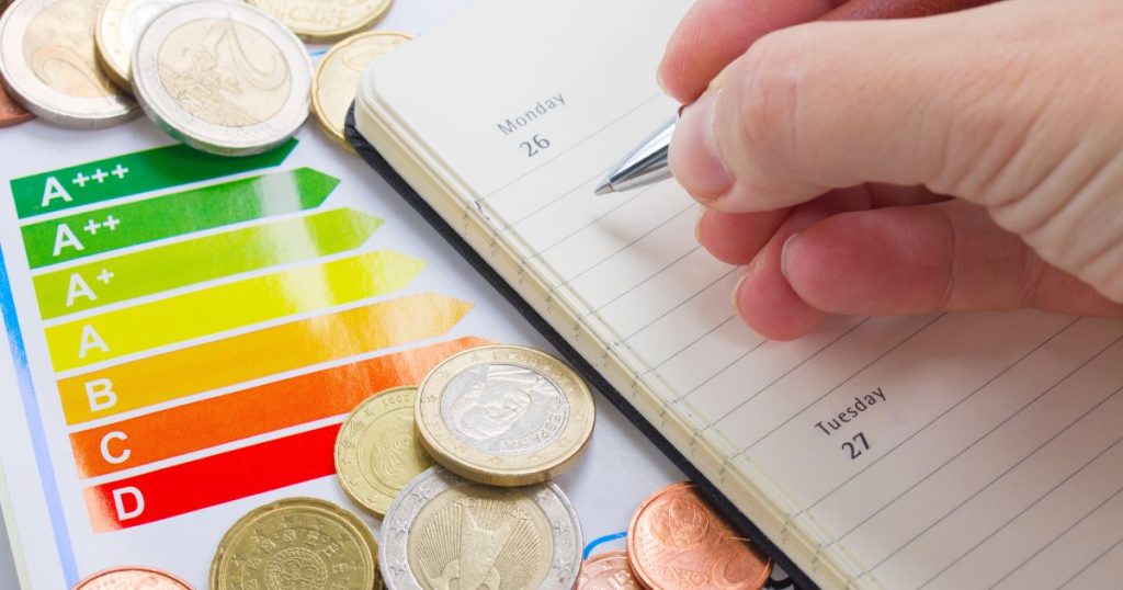 Een hand met pen boven een kleine agenda, bovenop een weergave van de EPC-scores bezaaid met euromunten en centen.